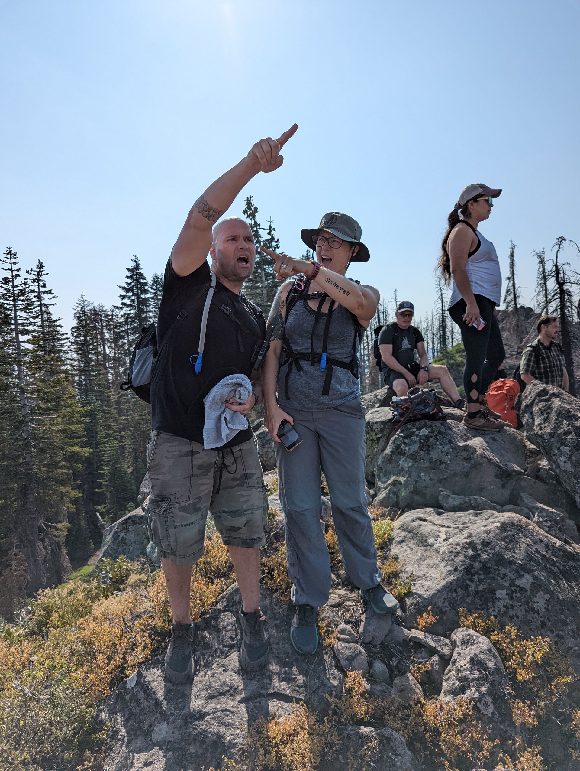 People hiking on a rocky mountain, man pointing upward, woman looking at his direction. Others in the background. Sunny day.