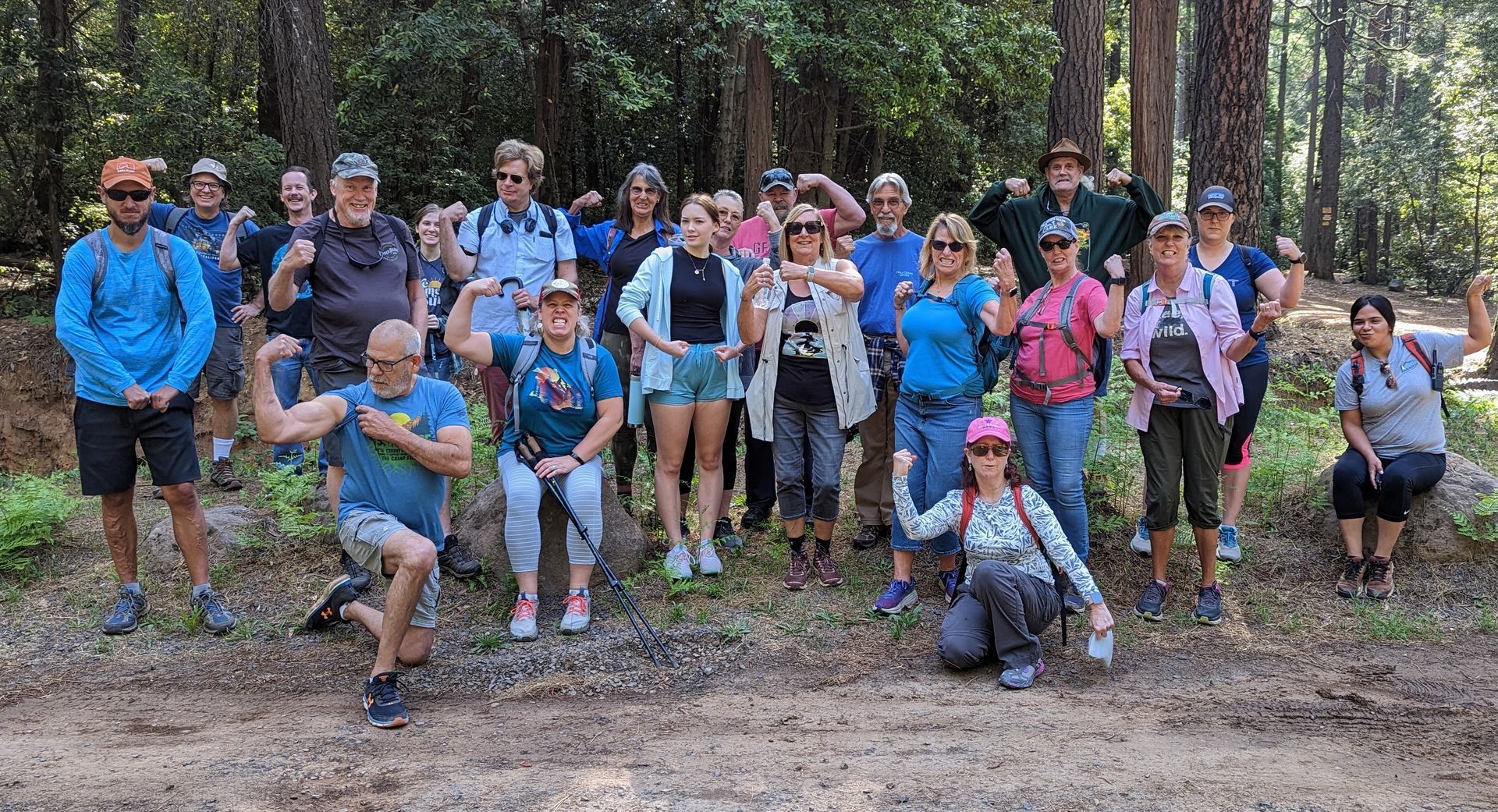 Group of people posing outdoors, many flexing muscles. Forest setting with trees in background.