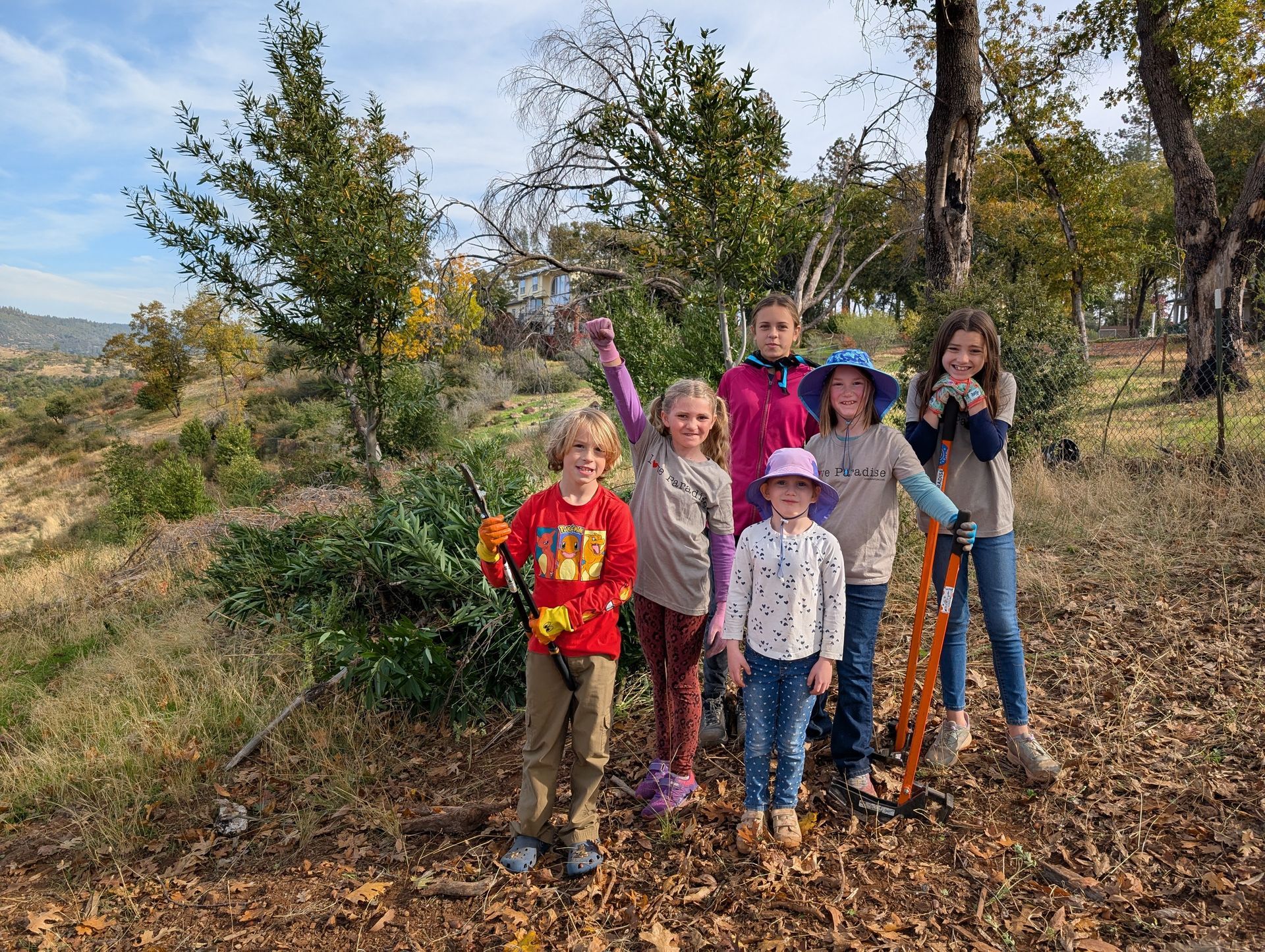 Group of children outdoors, smiling. One points up, others hold tools, fall leaves, trees, blue sky.