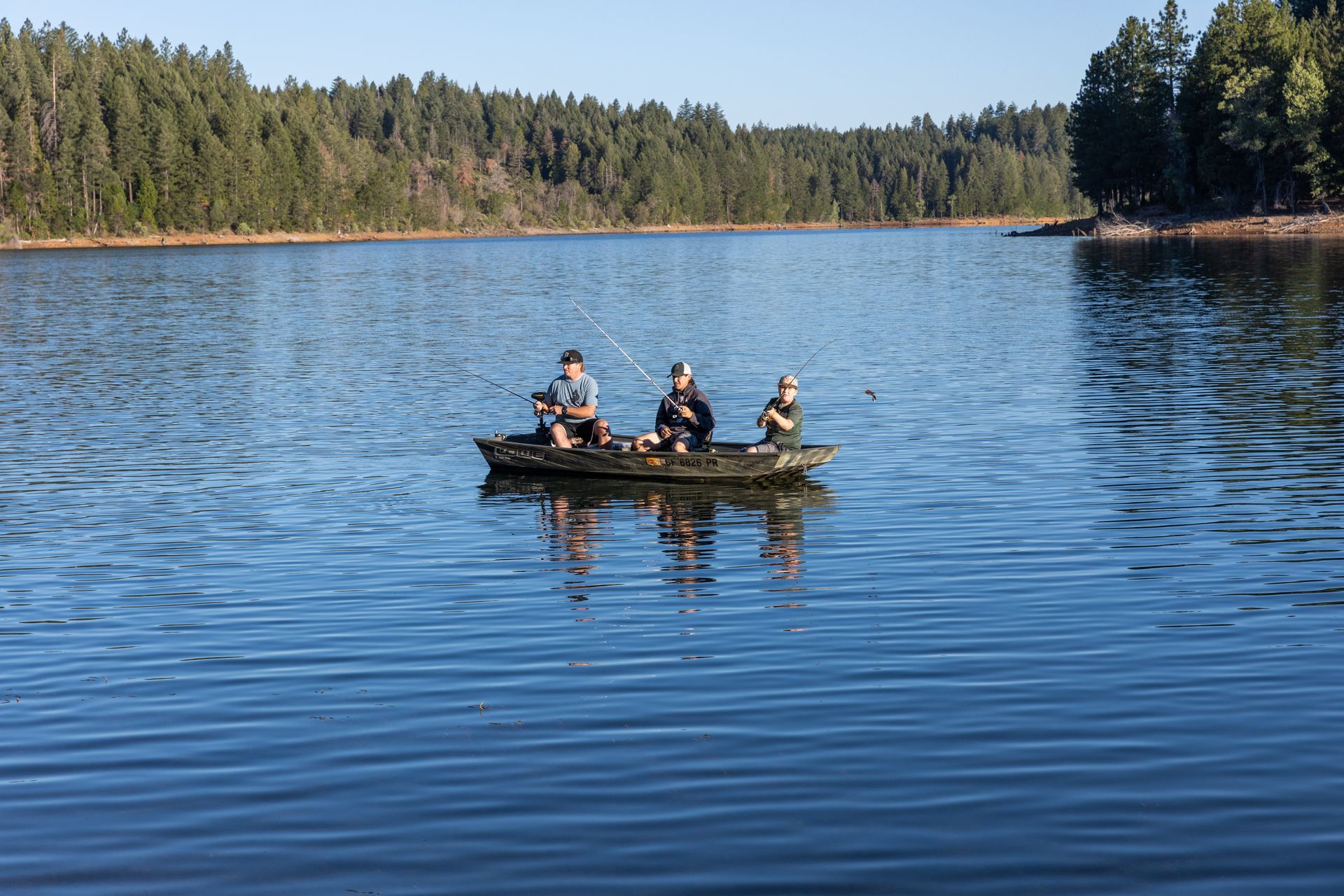 People fishing from a boat on a calm lake, surrounded by trees.