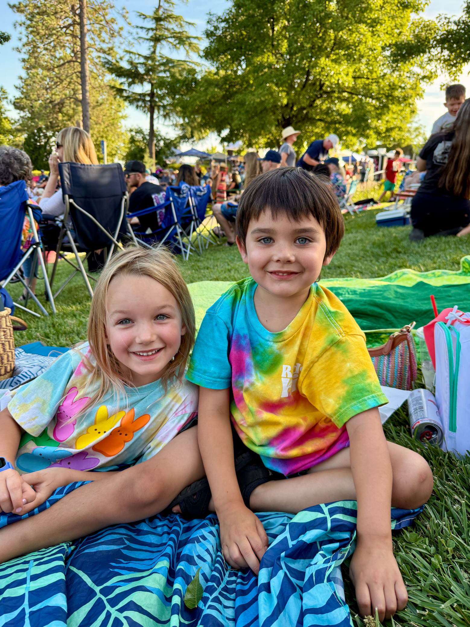 Two children in colorful tie-dye shirts sit on a blanket at an outdoor picnic, smiling at the camera.