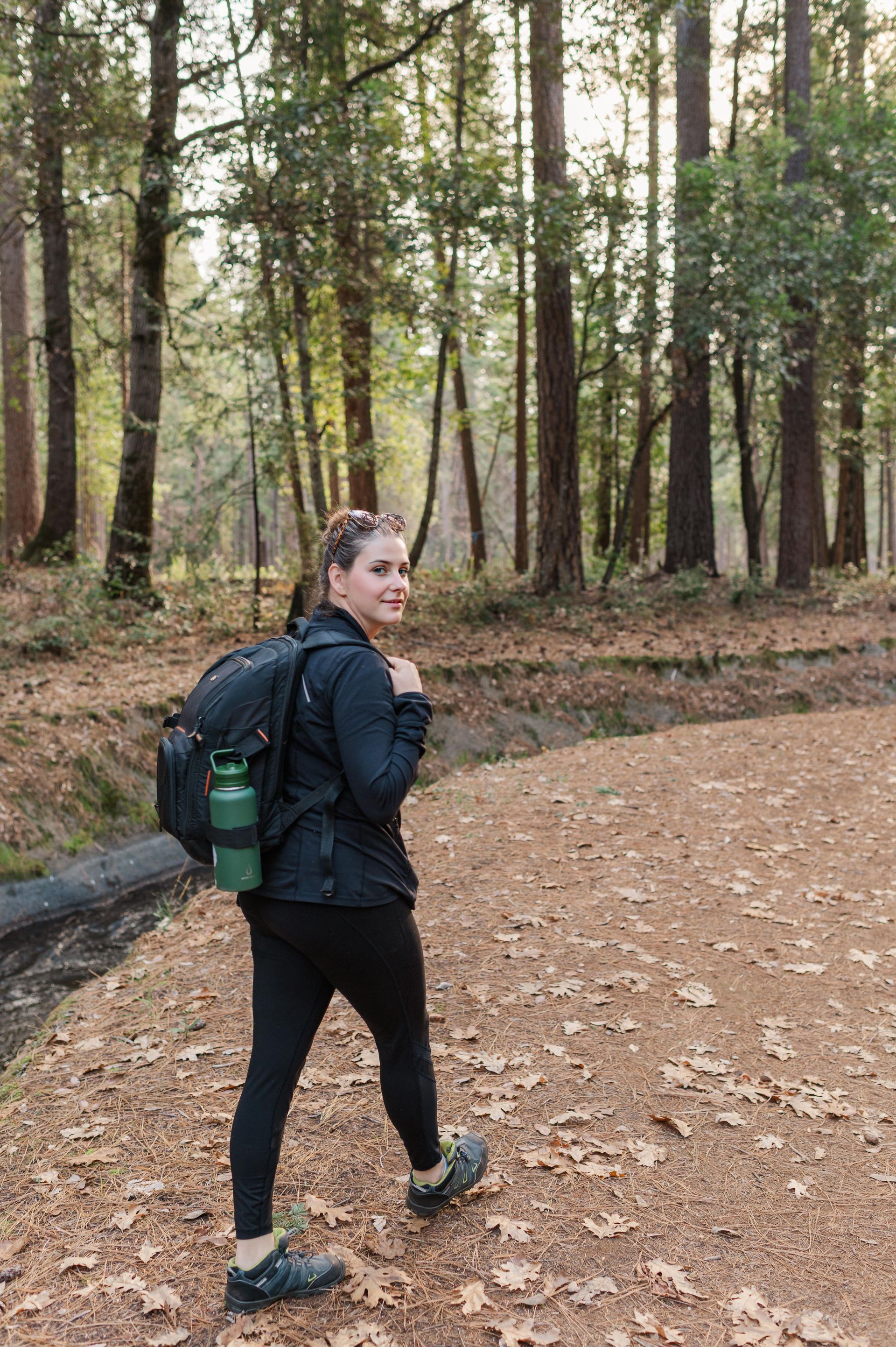 Woman hikes through a forest, wearing black clothing and a backpack with water bottle.