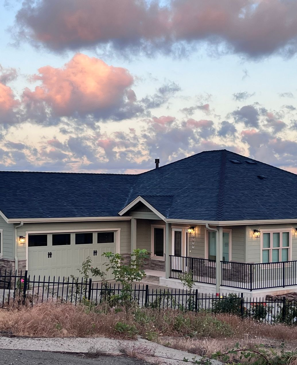 Green house with dark roof against a cloudy sky. Black fence and garage door visible.