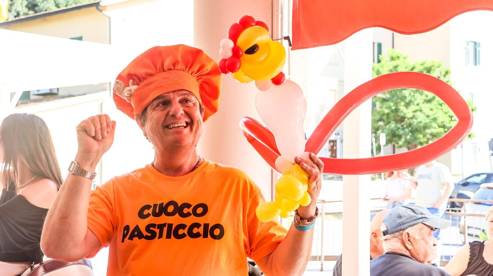 Un uomo che indossa una maglietta arancione con la scritta cuoco pasticcio tiene in mano dei palloncini.