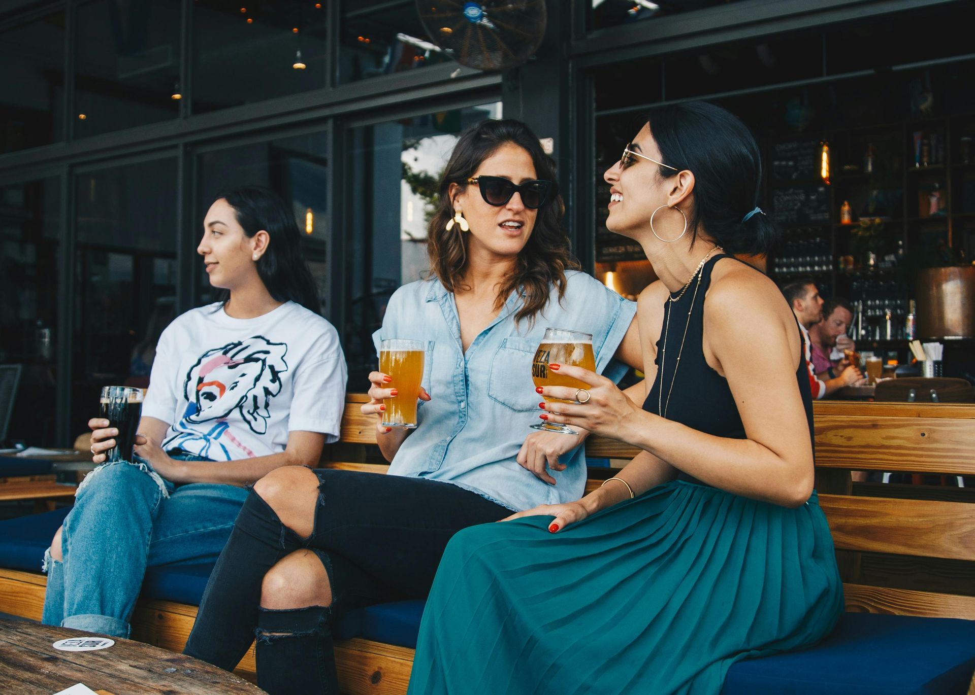 Three friends on a patio, holding beers. One wears sunglasses, laughing; others are talking. Wooden bench, bar in background.