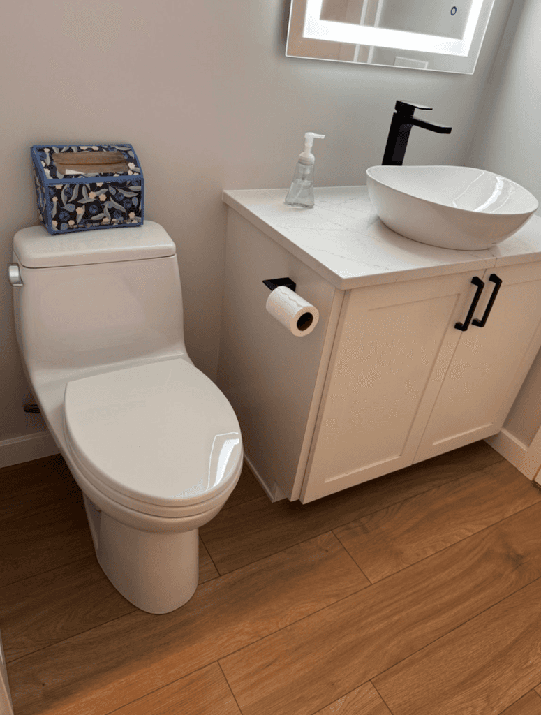 A small bathroom with a white toilet, white vanity with black handles, and a vessel sink. 