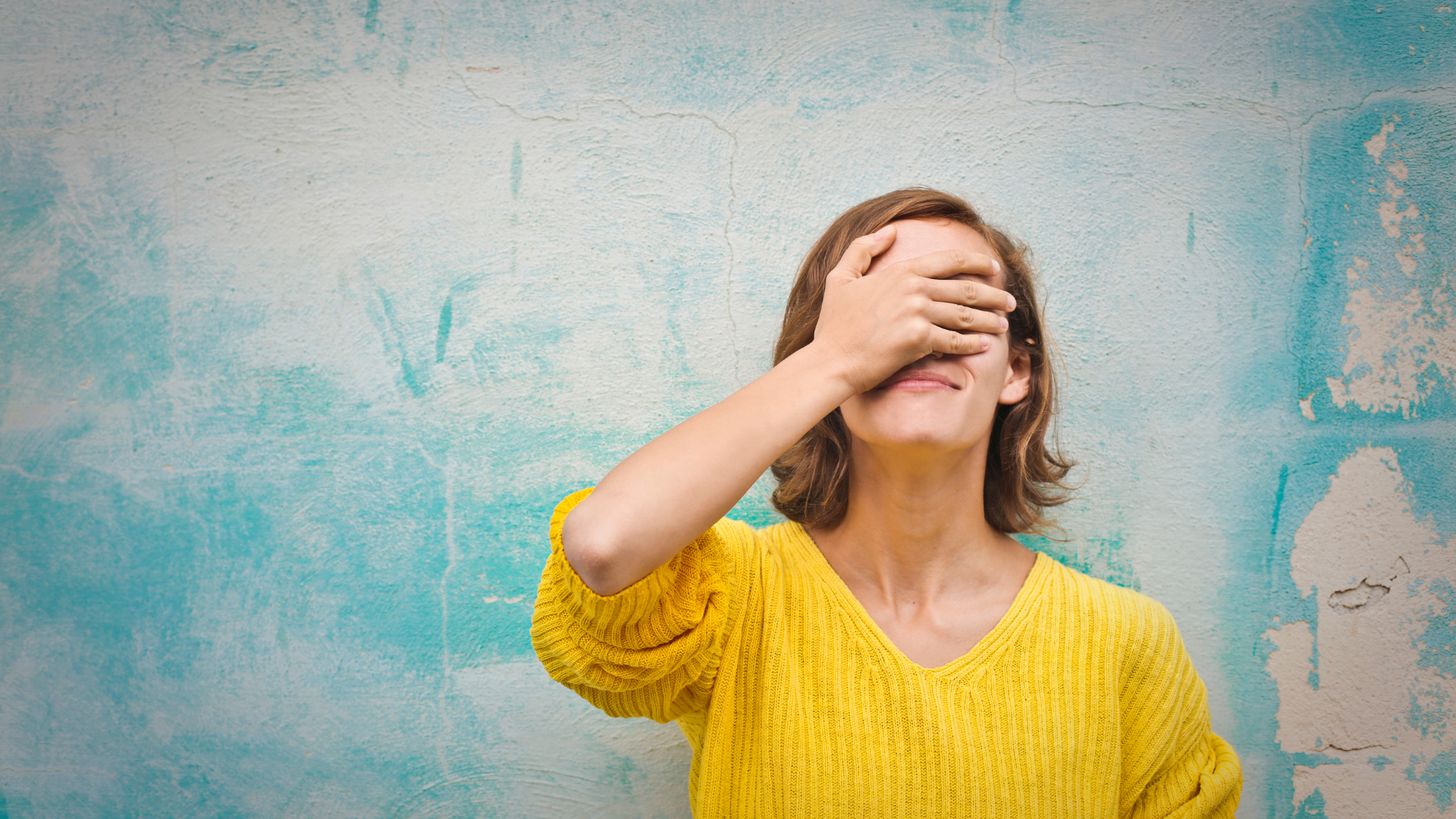 Person in a yellow sweater covering their face against a turquoise wall.