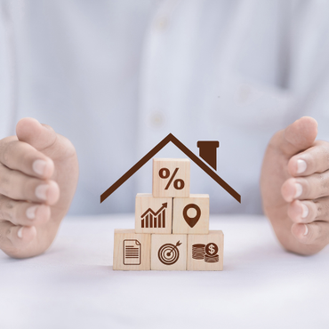 Hands framing small house-shaped wooden blocks with finance icons on a white background
