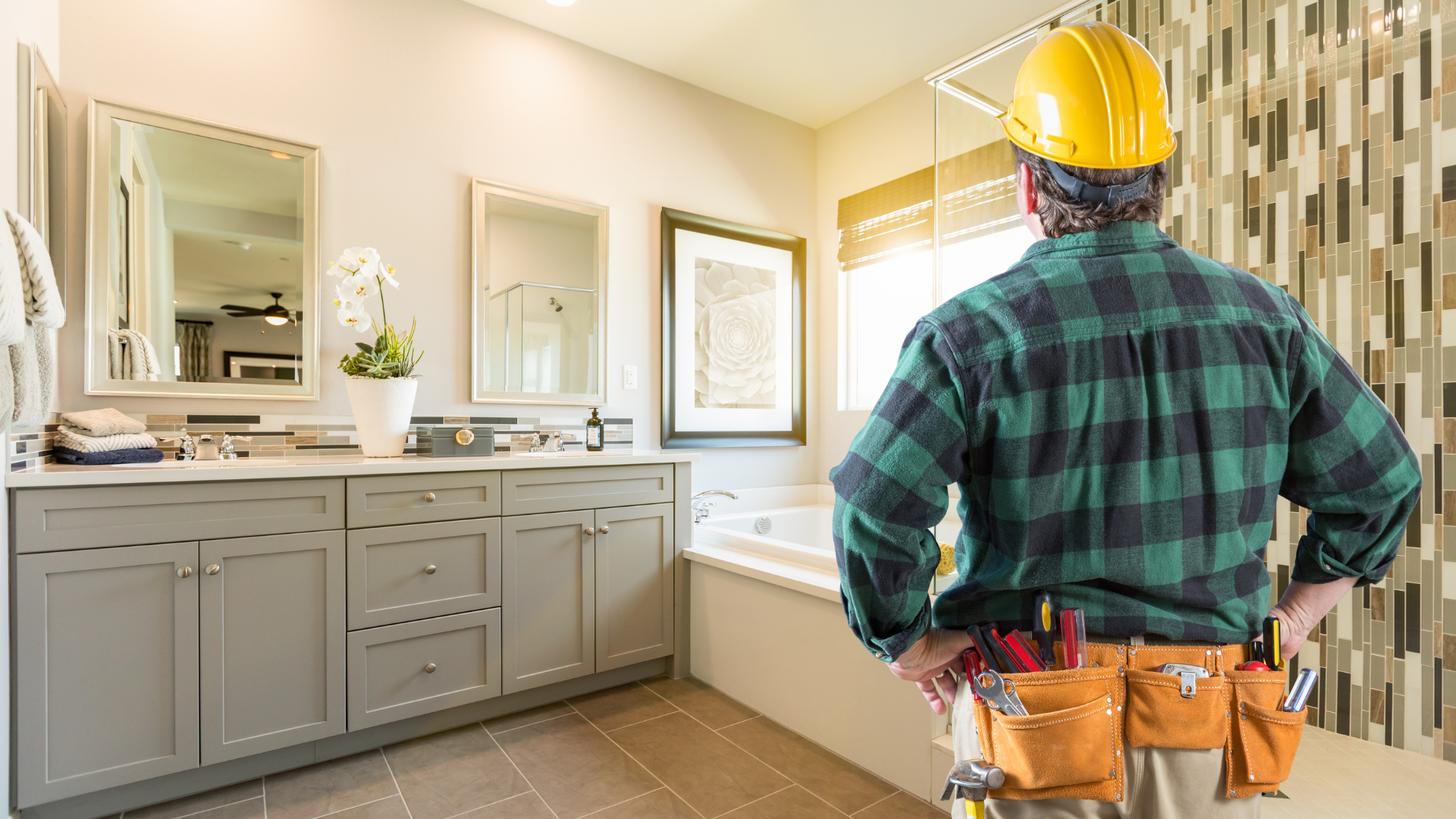 Contractor in hard hat standing in a bright bathroom with gray cabinets and mirrors