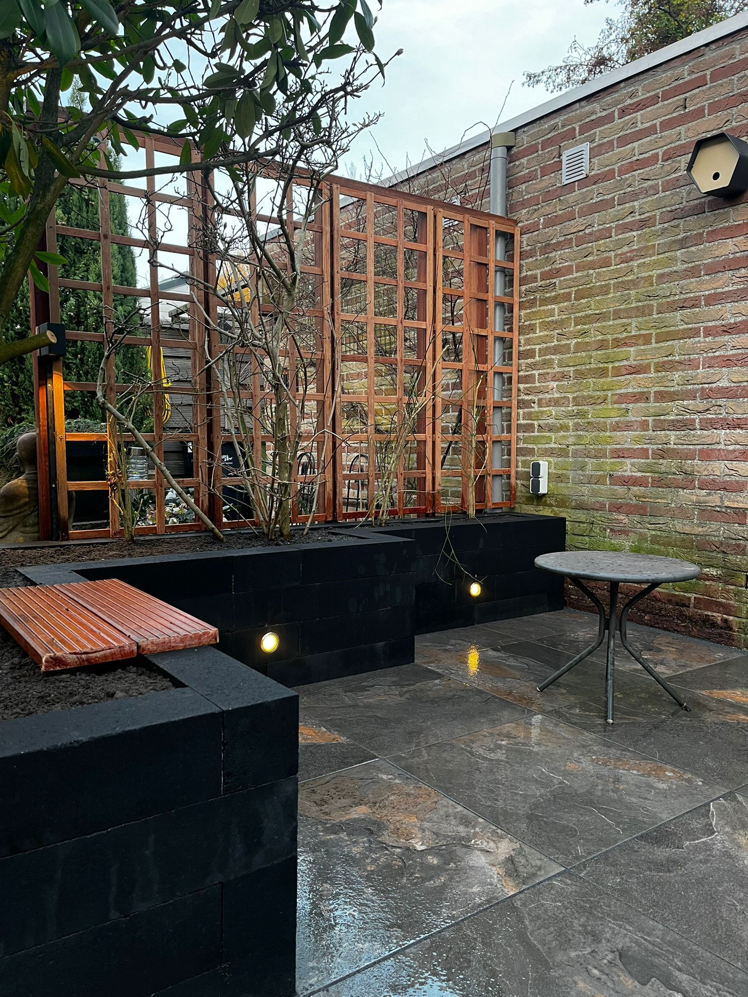 Buitenpatio met pergola, bakstenen muur en een klein tafeltje. Zwarte plantenbakken met ingebouwde verlichting.