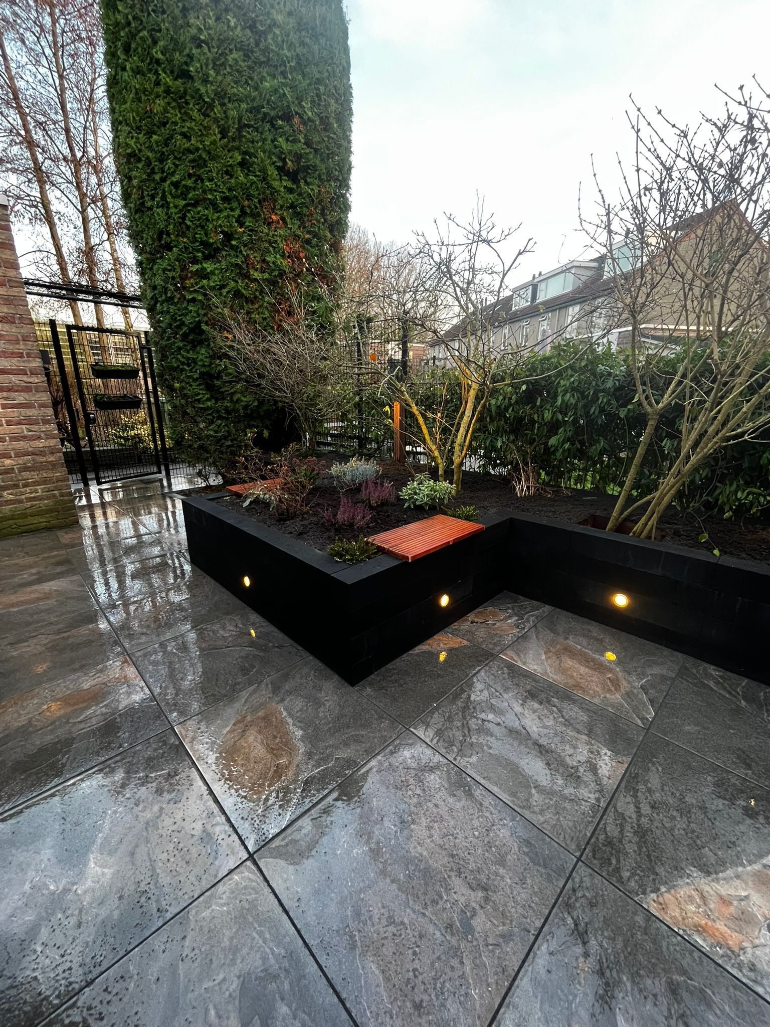 Donkere, moderne tuin met een terras van natuursteen, een zwarte plantenbak en ingebouwde verlichting.