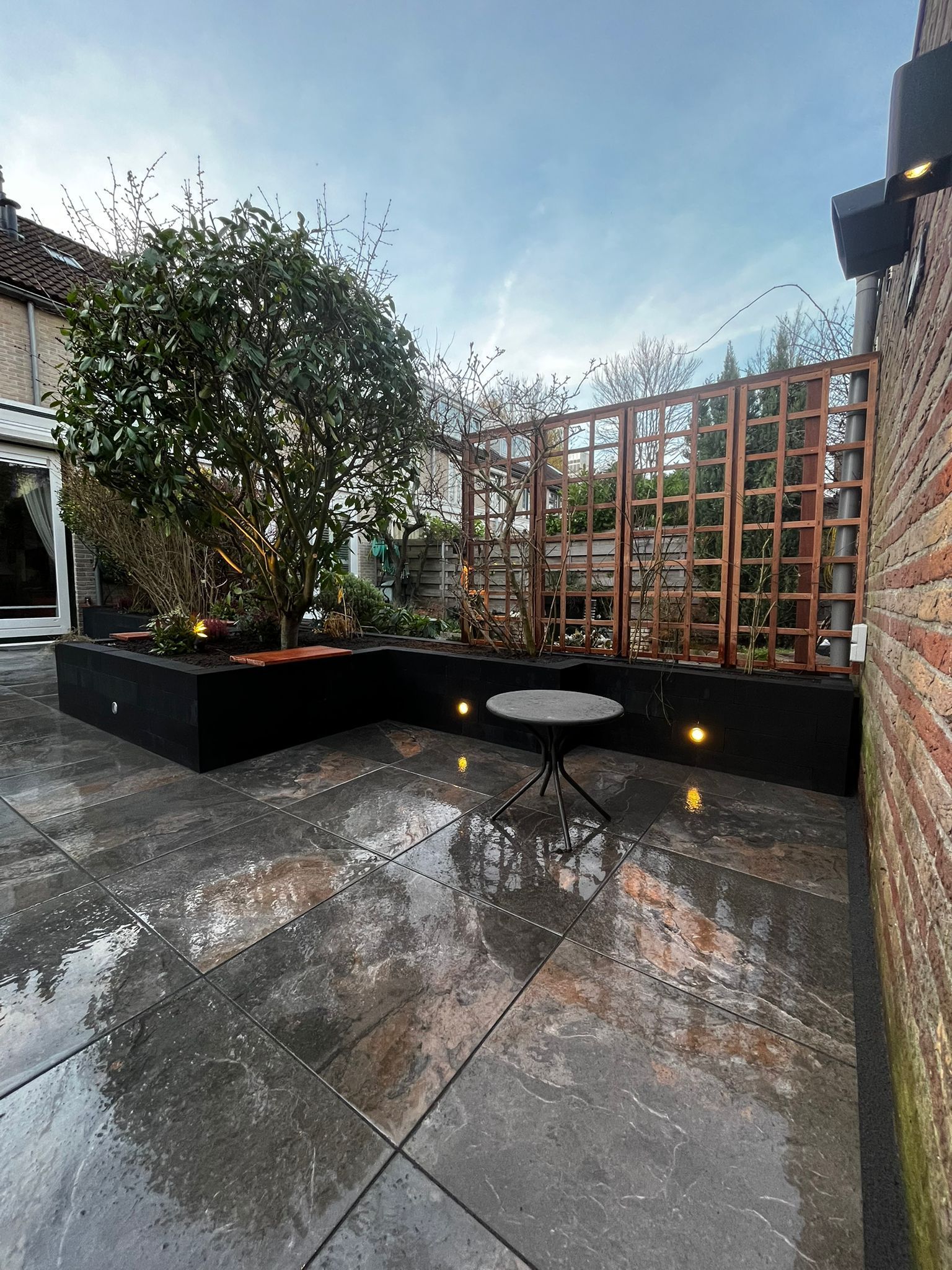 Terras met een zwarte plantenbak, houten pergola, klein tafeltje en een grote boom, die de natte grond weerspiegelt.