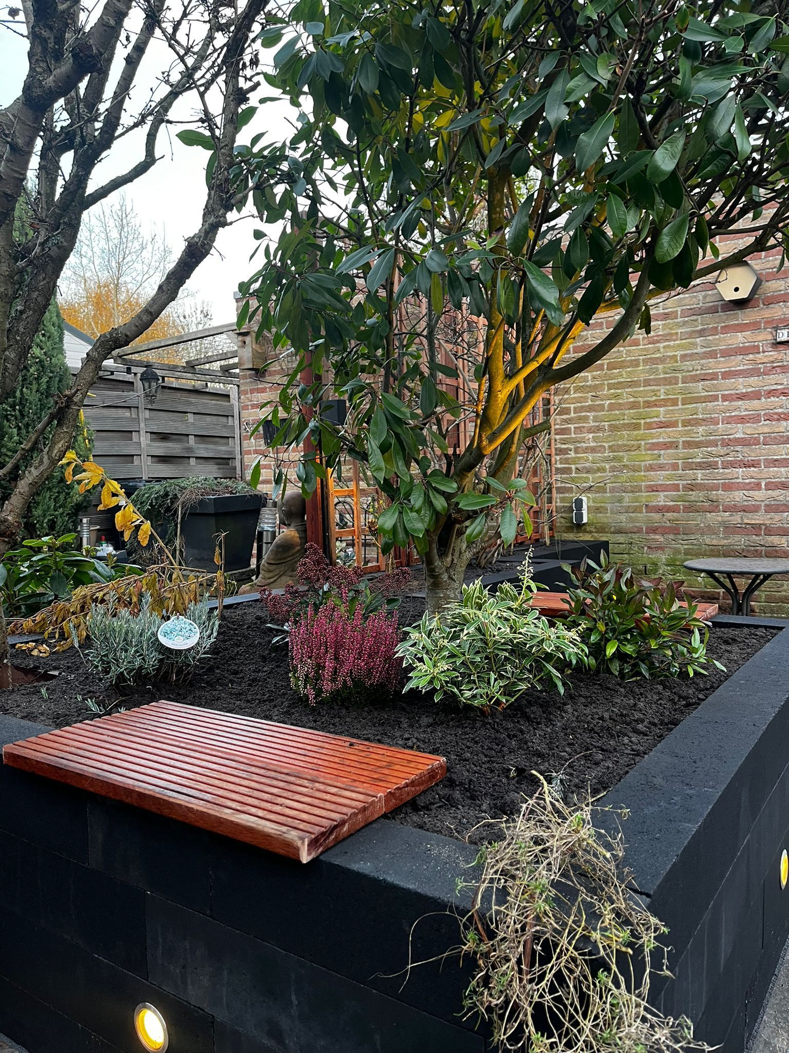 Een verhoogd bloembed met diverse planten en een houten bank voor een bakstenen gebouw.
