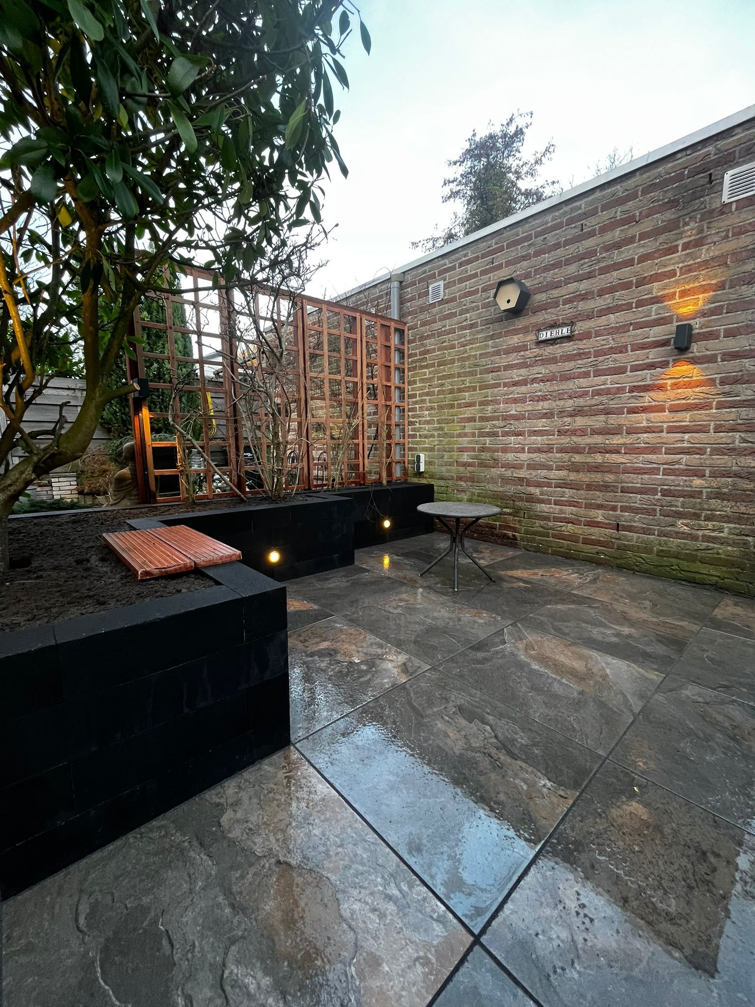 Terras met zwarte plantenbakken, stenen bestrating, bakstenen muur en decoratieve houten constructie.