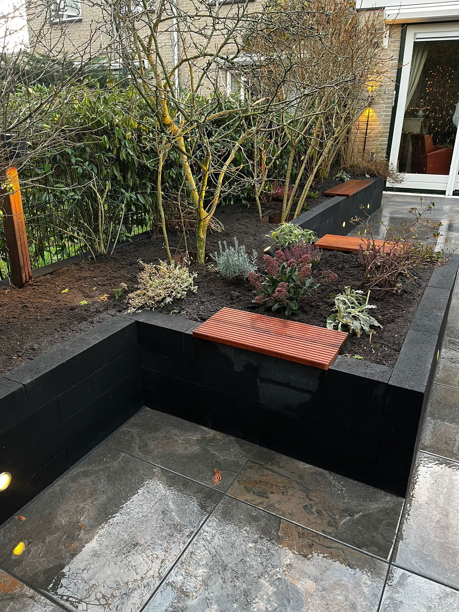 Verhoogd tuinbed met donkere rand, planten en een vierkant stenen terras.