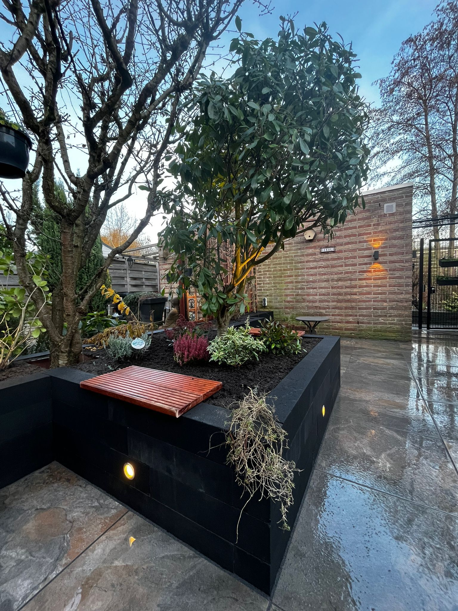 Zwarte plantenbak met bloemen, bomen en kleine lampjes in een tuinomgeving.