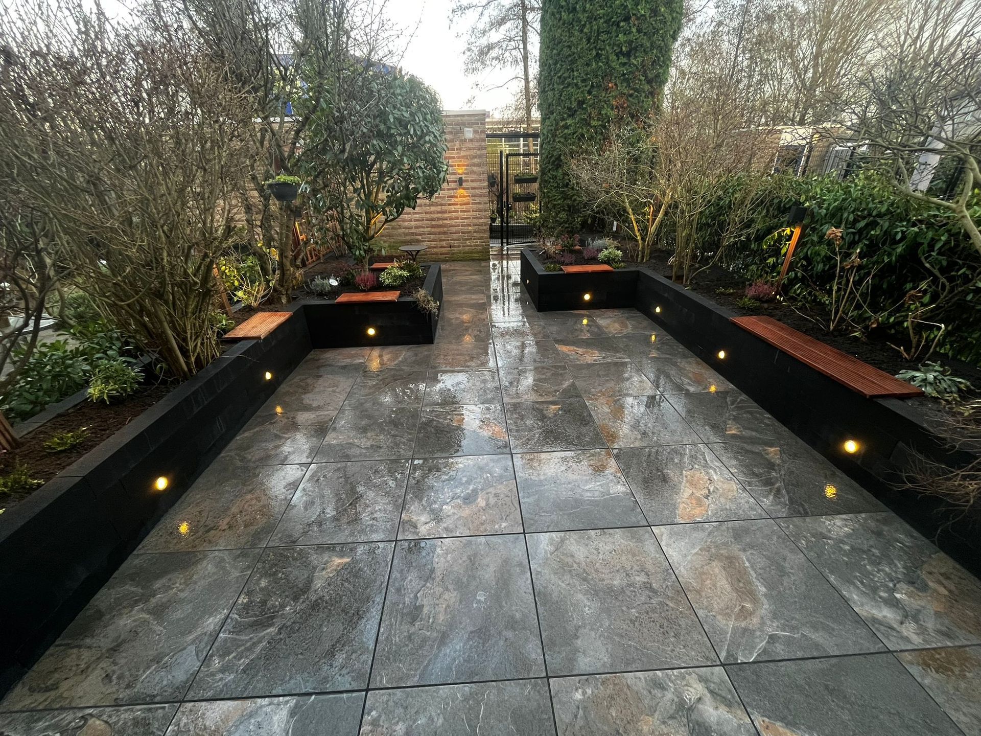 Stenen terras met ingebouwde plantenbakken en verlichting, dat leidt naar een trap en een poort, geflankeerd door struiken.