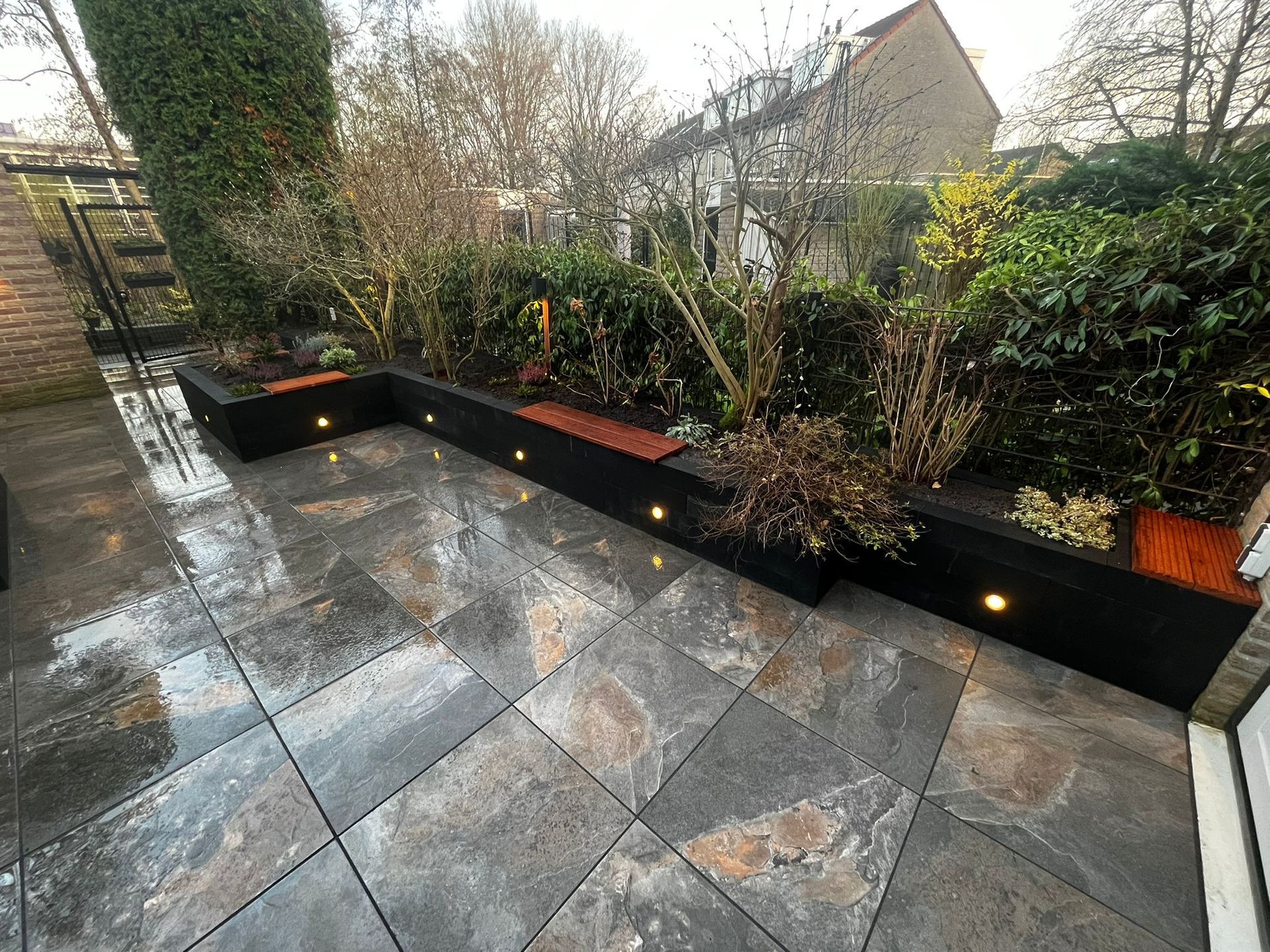 Zwarte plantenbakken met ingebouwde verlichting op een terras van natuursteen, omgeven door hagen en een schutting.