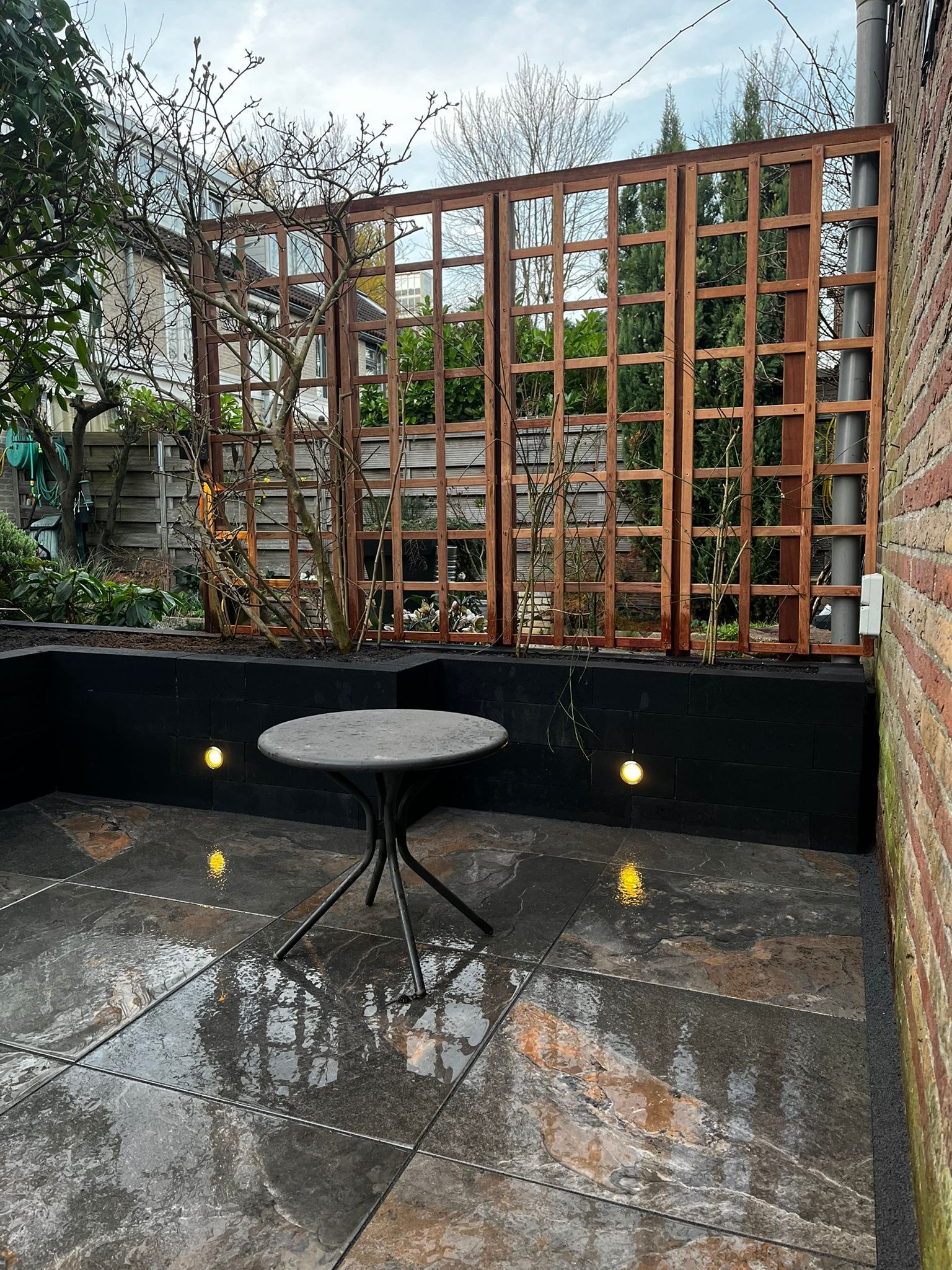 Terras met een klein tafeltje, een pergola en inbouwspots. Rechts een bakstenen muur. Het natte wegdek weerkaatst het licht.