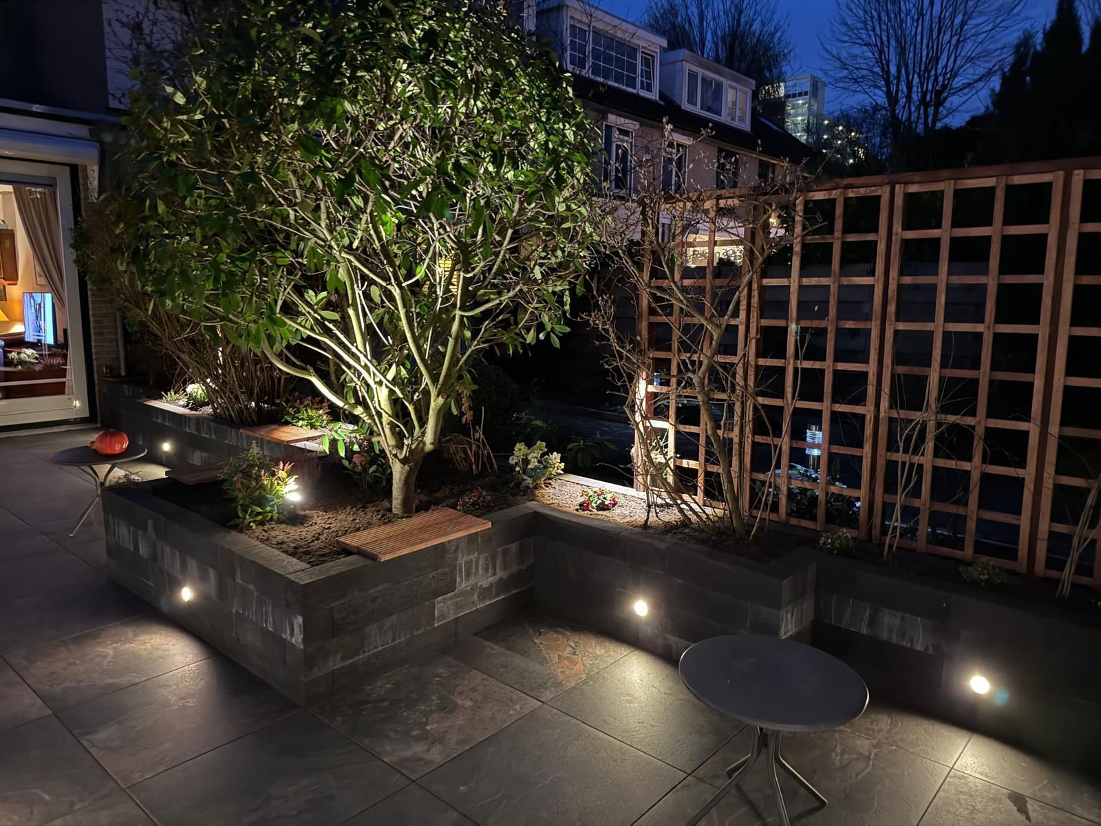 Terras 's avonds met verlichte boom, plantenbakken, pergola en tafel. Donkergrijze steen, warme verlichting.
