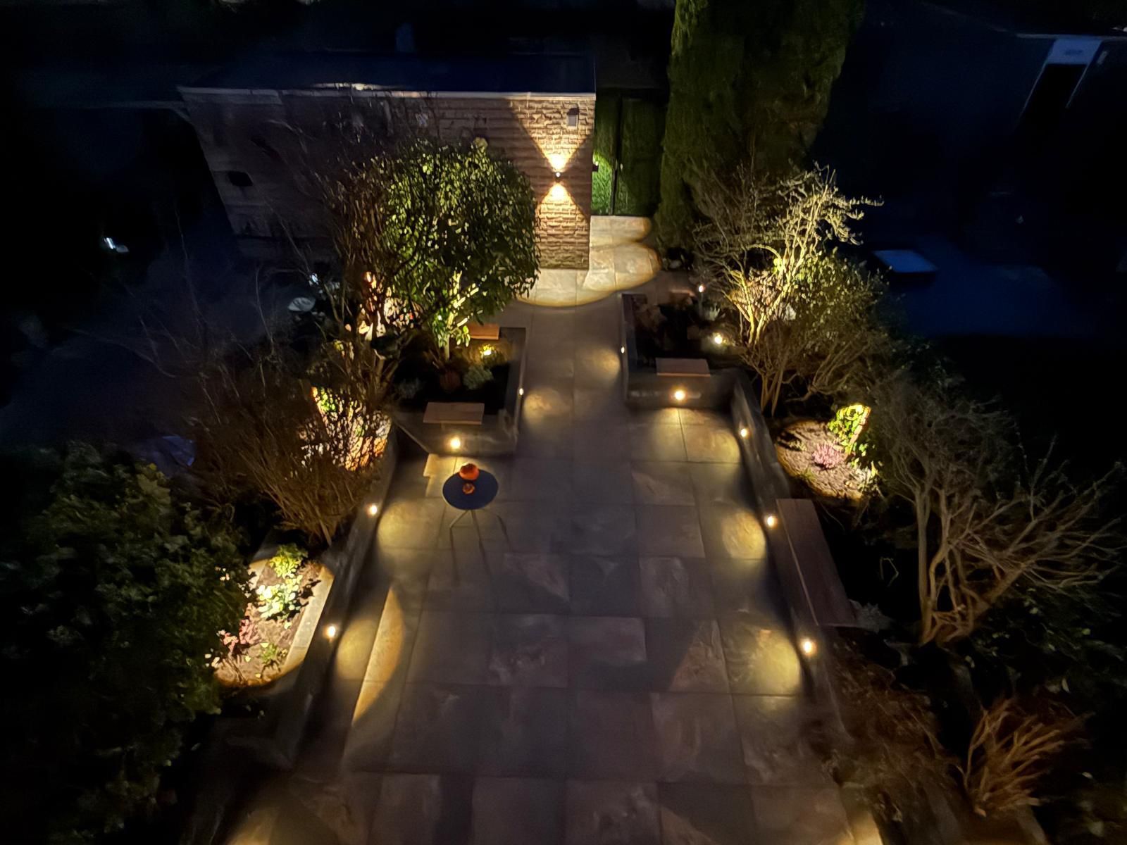 Een nachtopname van een verlicht terraspad met planten en bomen. Een schijnwerper verlicht het pad, de hagen en een houten constructie.