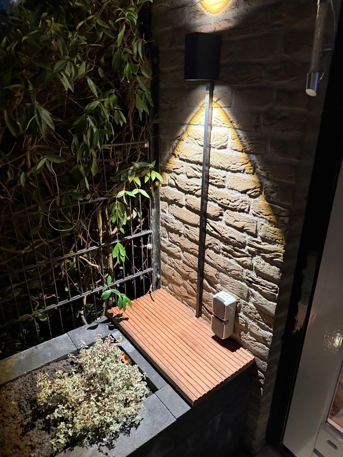 Buitenhoek met bakstenen muur, houten terras, kleine tuin en 's nachts verlicht door een spot.