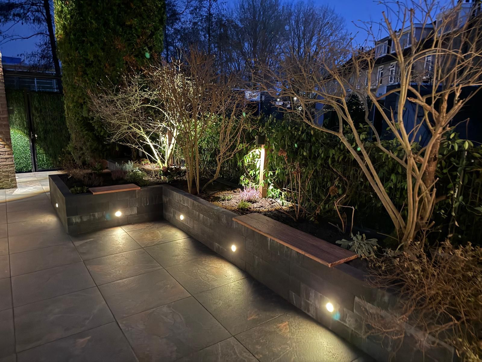 Nachtelijk uitzicht op een patio met ingebouwde plantenbakken en tuinverlichting. De stenen patio en de tuinmuur zijn verlicht.