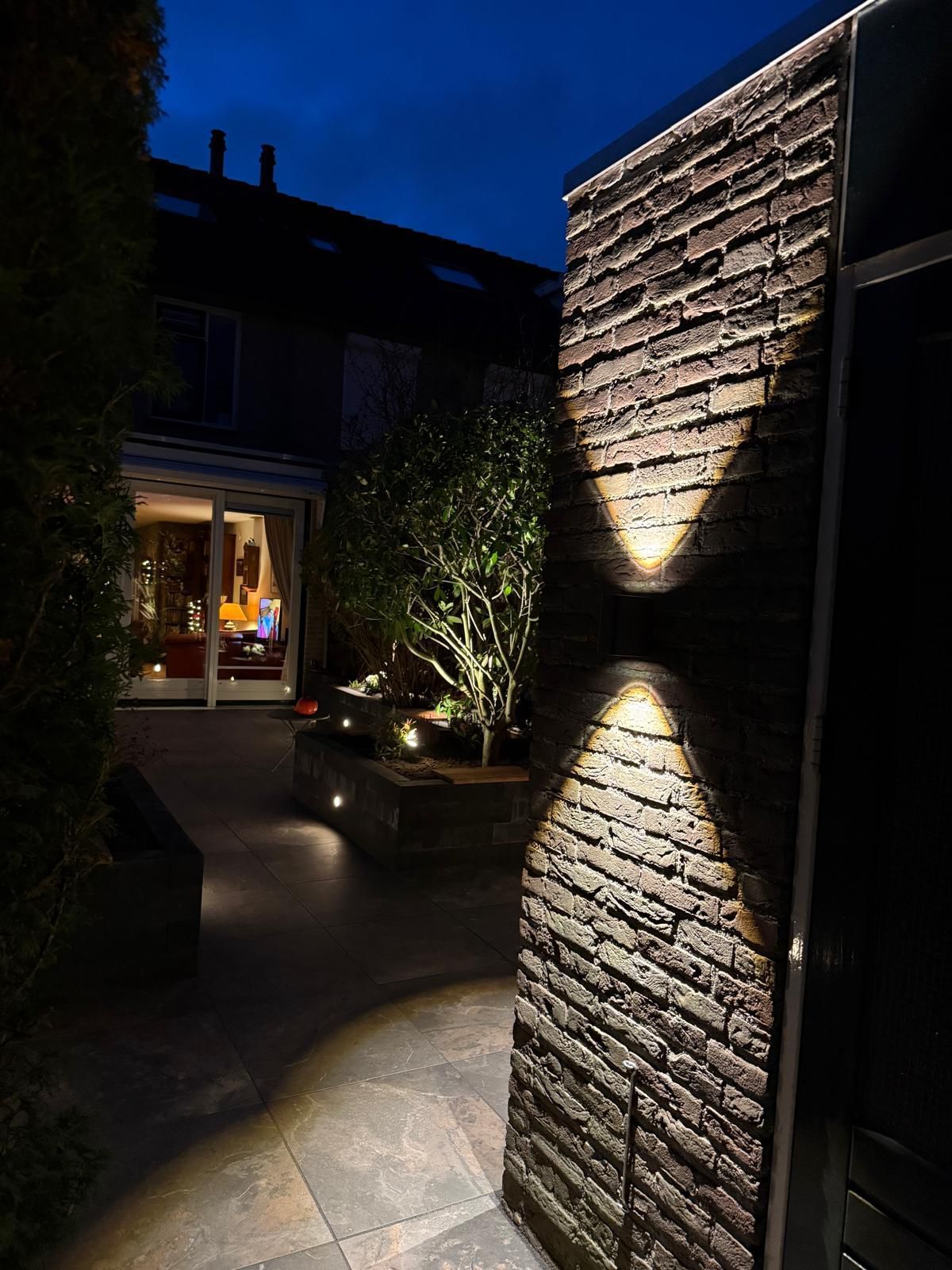 Buitenruimte 's nachts, verlichte bakstenen muur met up/down-verlichting, donkerblauwe lucht, terras, huis op de achtergrond.