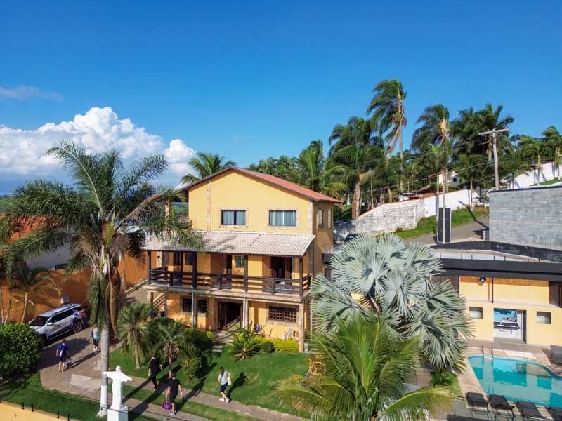 Casa de praia amarela com varanda, palmeiras e piscina. Céu azul ao fundo.