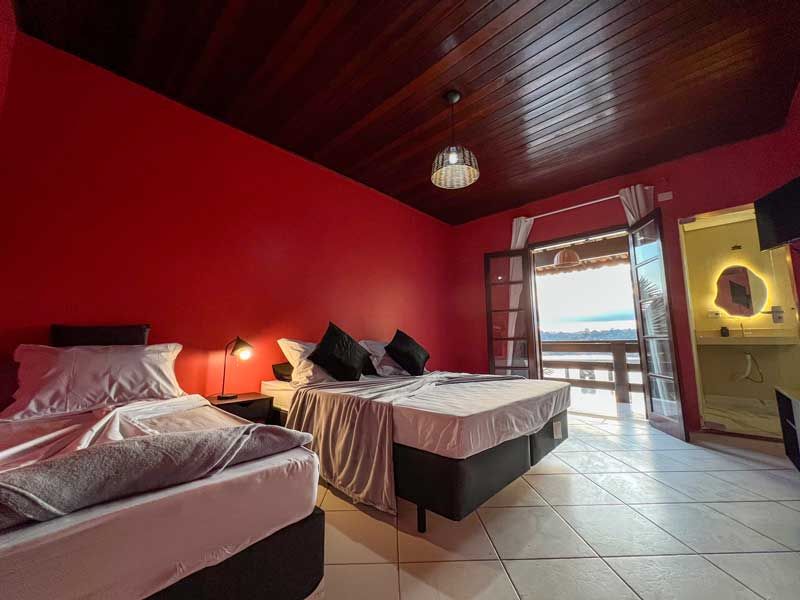 Quarto com paredes vermelhas, três camas e vista para a varanda.
