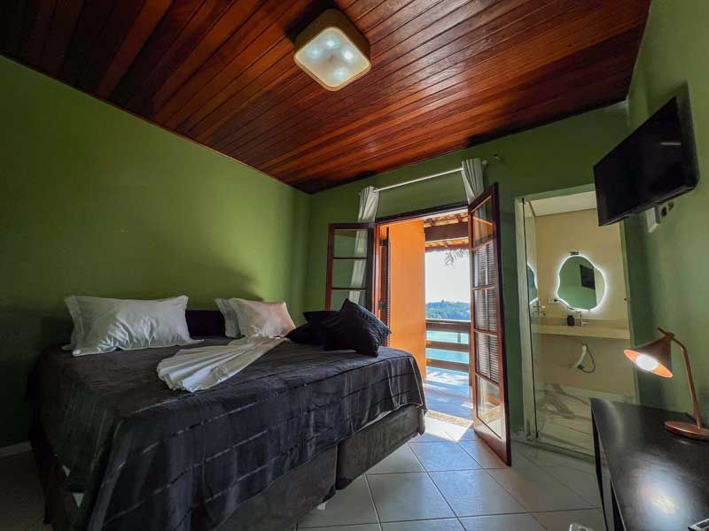 Quarto com paredes verdes, cama escura, teto de madeira, portas abertas com vista e banheiro pequeno.