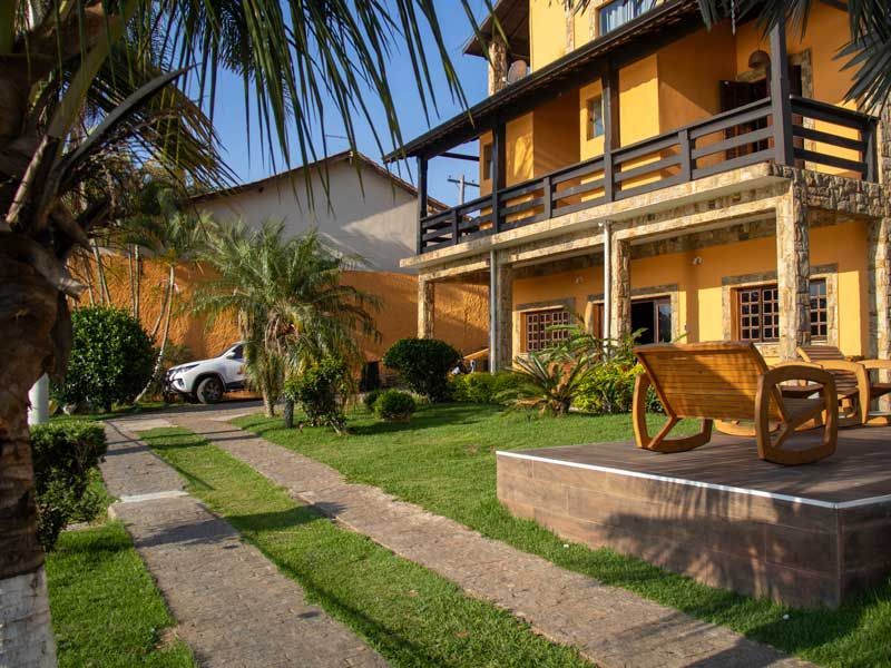 Casa amarela de dois andares com varanda, gramado, palmeiras e carro estacionado na entrada.