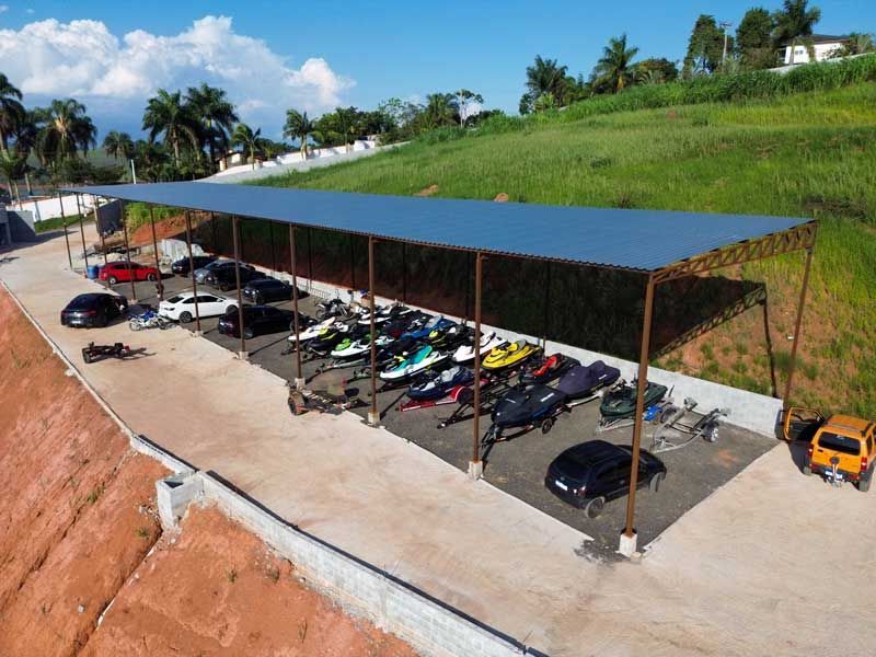 Carros e jet skis estacionados sob uma grande estrutura sombreada em um terreno de concreto, com uma colina gramada ao fundo.