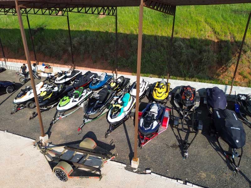 Jet skis em reboques estacionados sob uma cobertura. Alguns têm capas. Grama verde ao fundo.