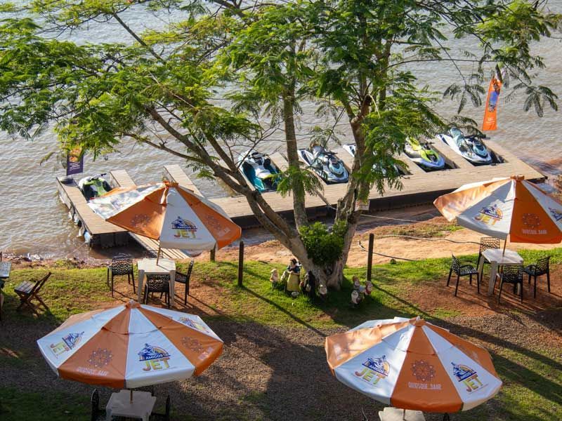 Uma cena à beira-mar com guarda-sóis laranja, mesas, jet skis em um píer e árvores.