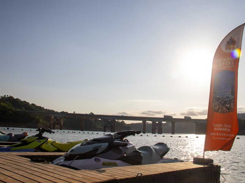 Jet skis atracados em um píer ao pôr do sol, ponte ao fundo, bandeira laranja.