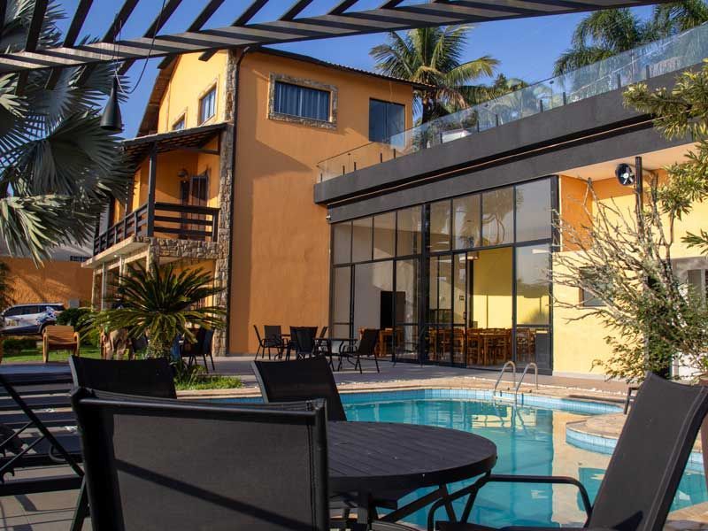 Área da piscina com um prédio de dois andares ao fundo. Mesas ao ar livre com pérgola coberta.