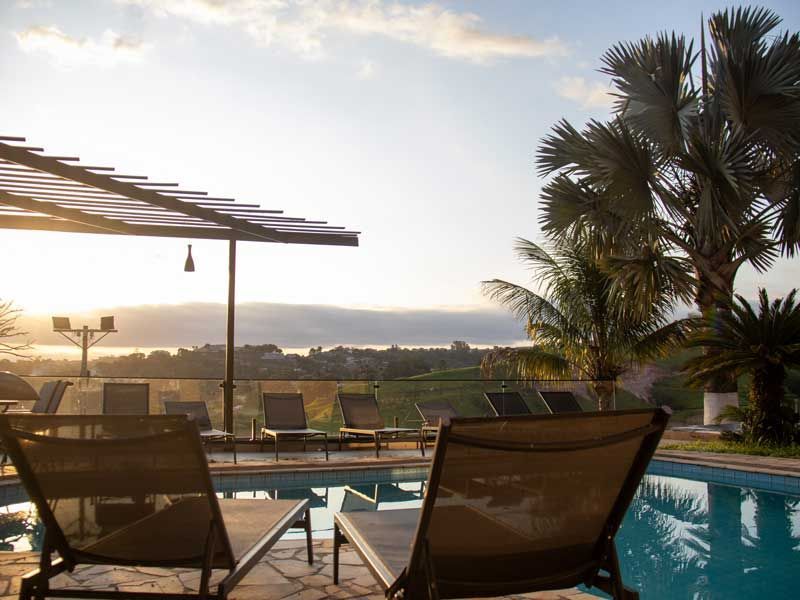 Vista da piscina ao nascer do sol com espreguiçadeiras, palmeiras e paisagem ao longe.