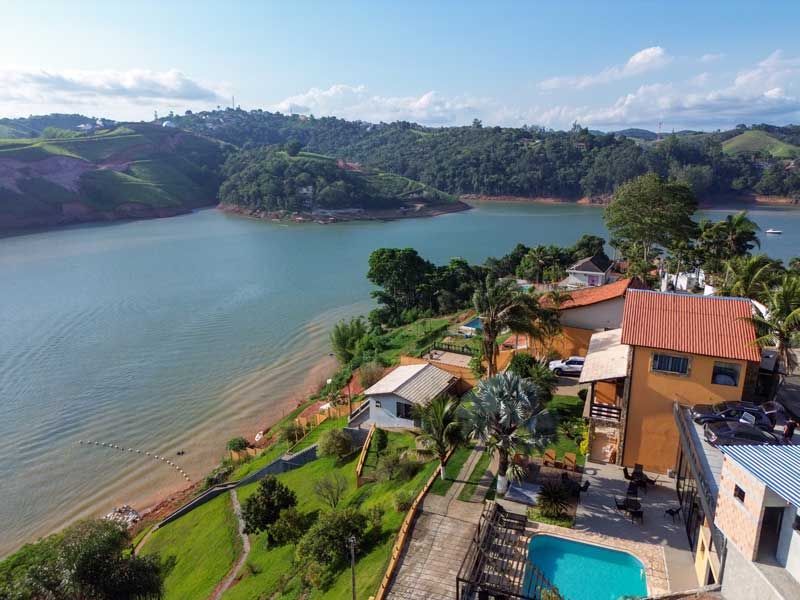 Casas e piscina perto de um lago, cercadas por colinas verdejantes sob um céu azul.