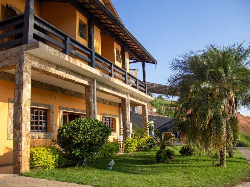 Casa amarela de dois andares com detalhes em pedra, uma varanda e um gramado bem cuidado com uma palmeira.