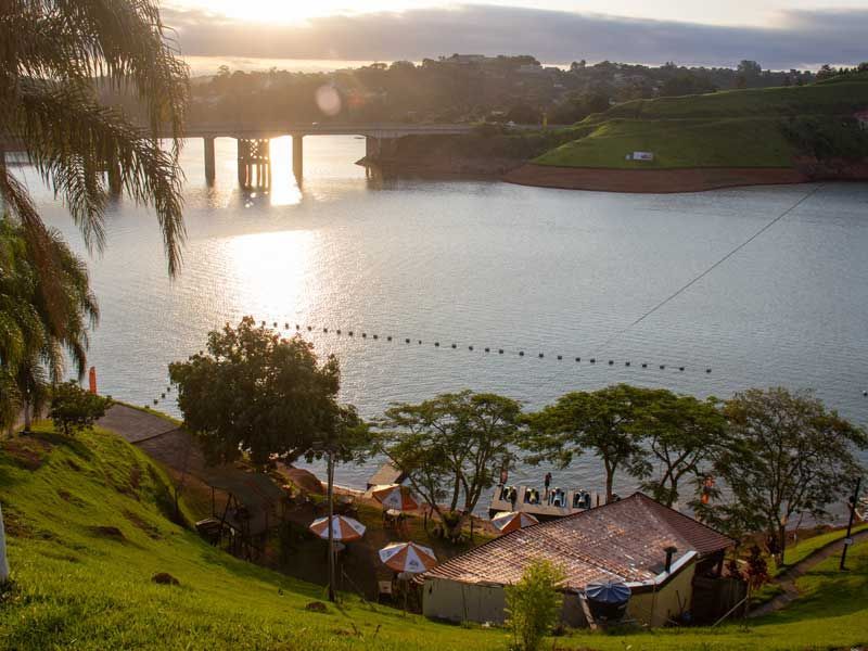 Cena tranquila de um lago ao pôr do sol, com uma ponte, um restaurante na margem e vegetação exuberante.