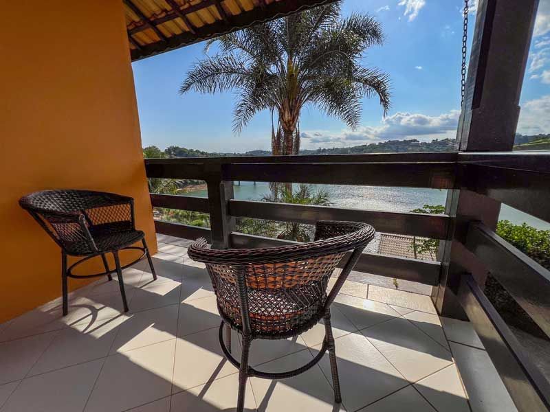 Duas cadeiras de vime em uma varanda com vista para um lago e uma palmeira. Dia ensolarado.