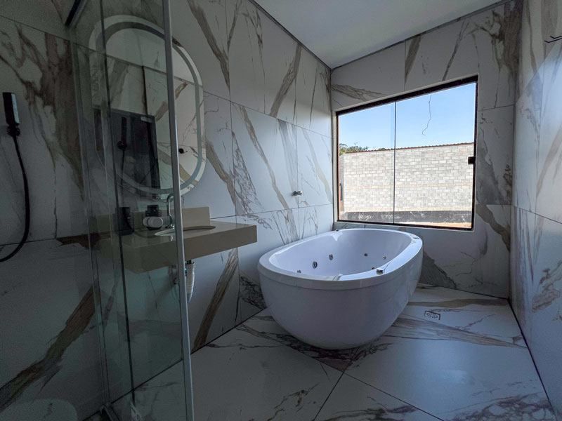 Banheiro moderno com banheira de hidromassagem branca, pia com bancada e paredes revestidas com azulejos de mármore. Uma janela oferece vista para o exterior.