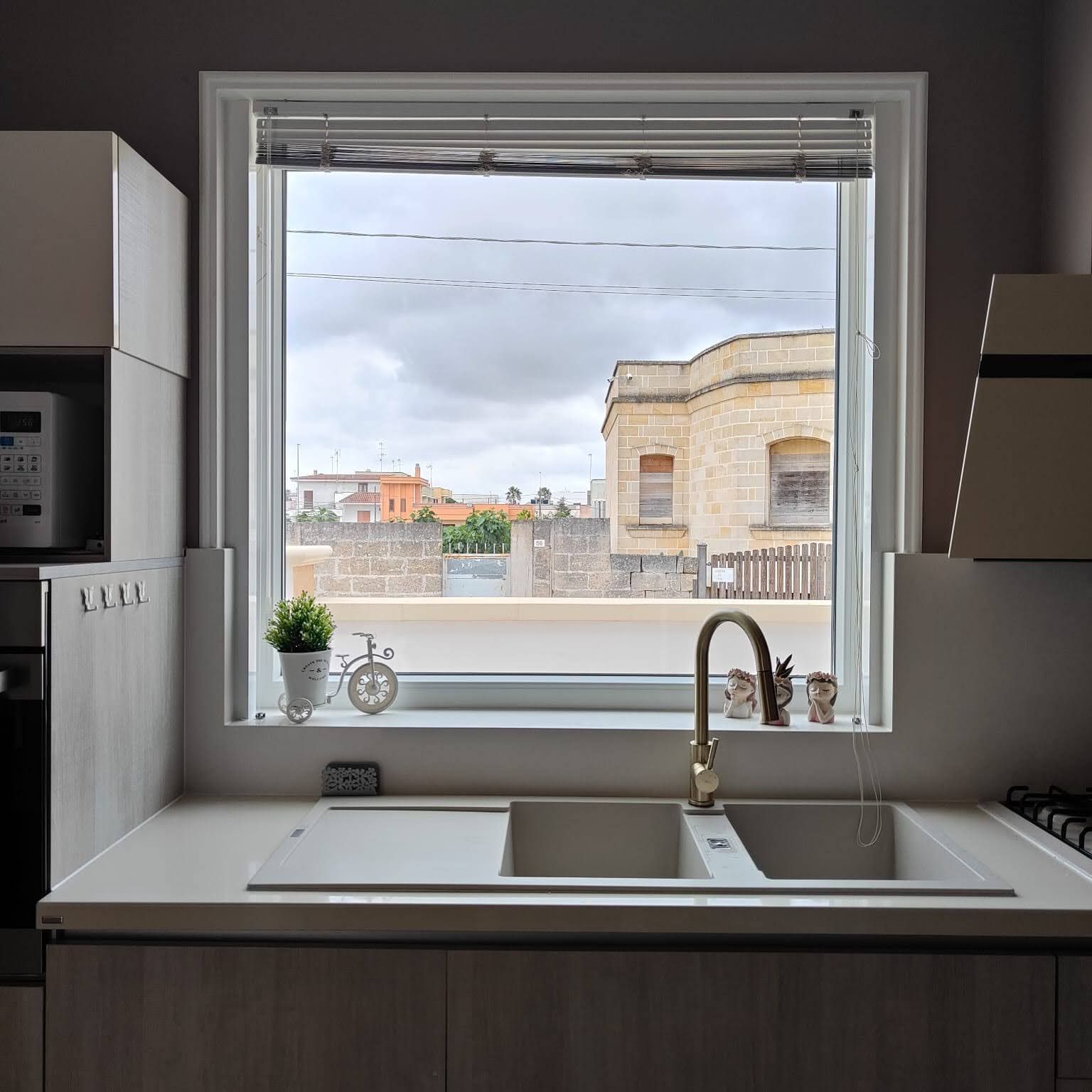 Un lavello da cucina con rubinetto dorato sotto una grande finestra che si affaccia su una strada fiancheggiata da edifici in pietra beige.