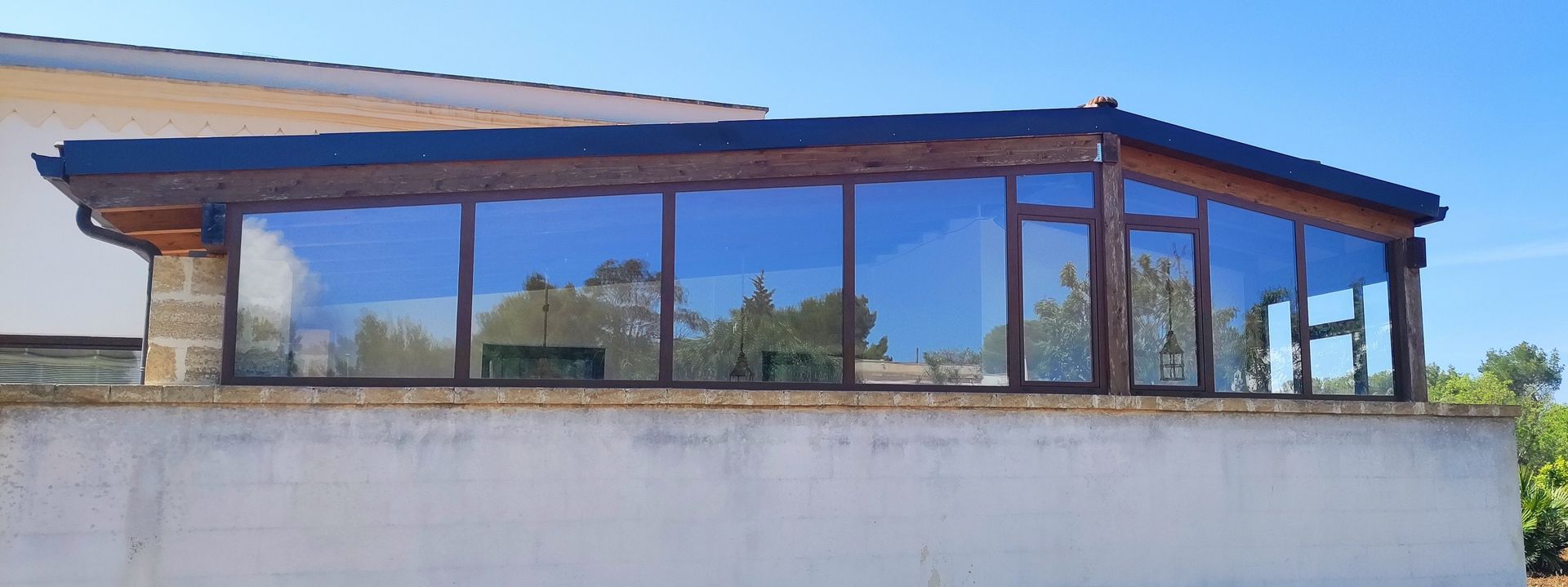 Una moderna veranda con ampie vetrate e una struttura in legno scuro, incastonata su una parete bianca sotto un cielo azzurro e limpido.