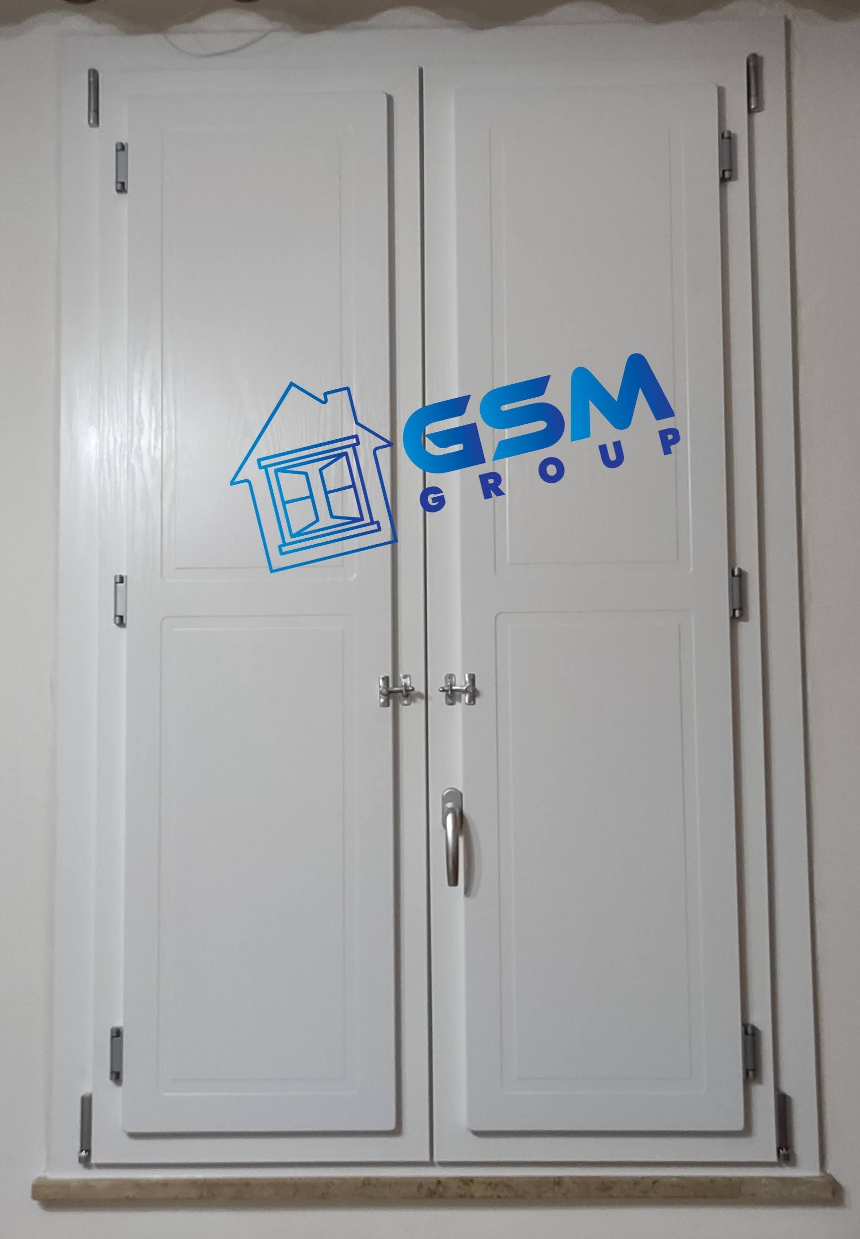Persiane interne bianche a doppio pannello installate su una parete, con un piccolo logo GSM Group sovrapposto al centro.