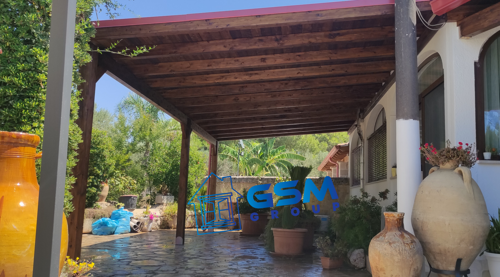 Un patio coperto con pergolato in legno, pavimento in pietra, grandi vasi in ceramica e rigogliose piante verdi in un ambiente da giardino.