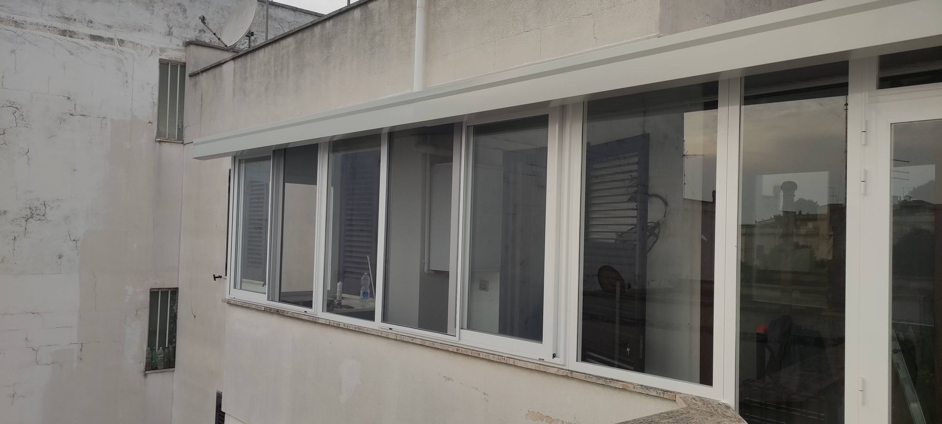 Una fila di finestre scorrevoli con cornice bianca installate sulla facciata esterna di un edificio beige con balcone.