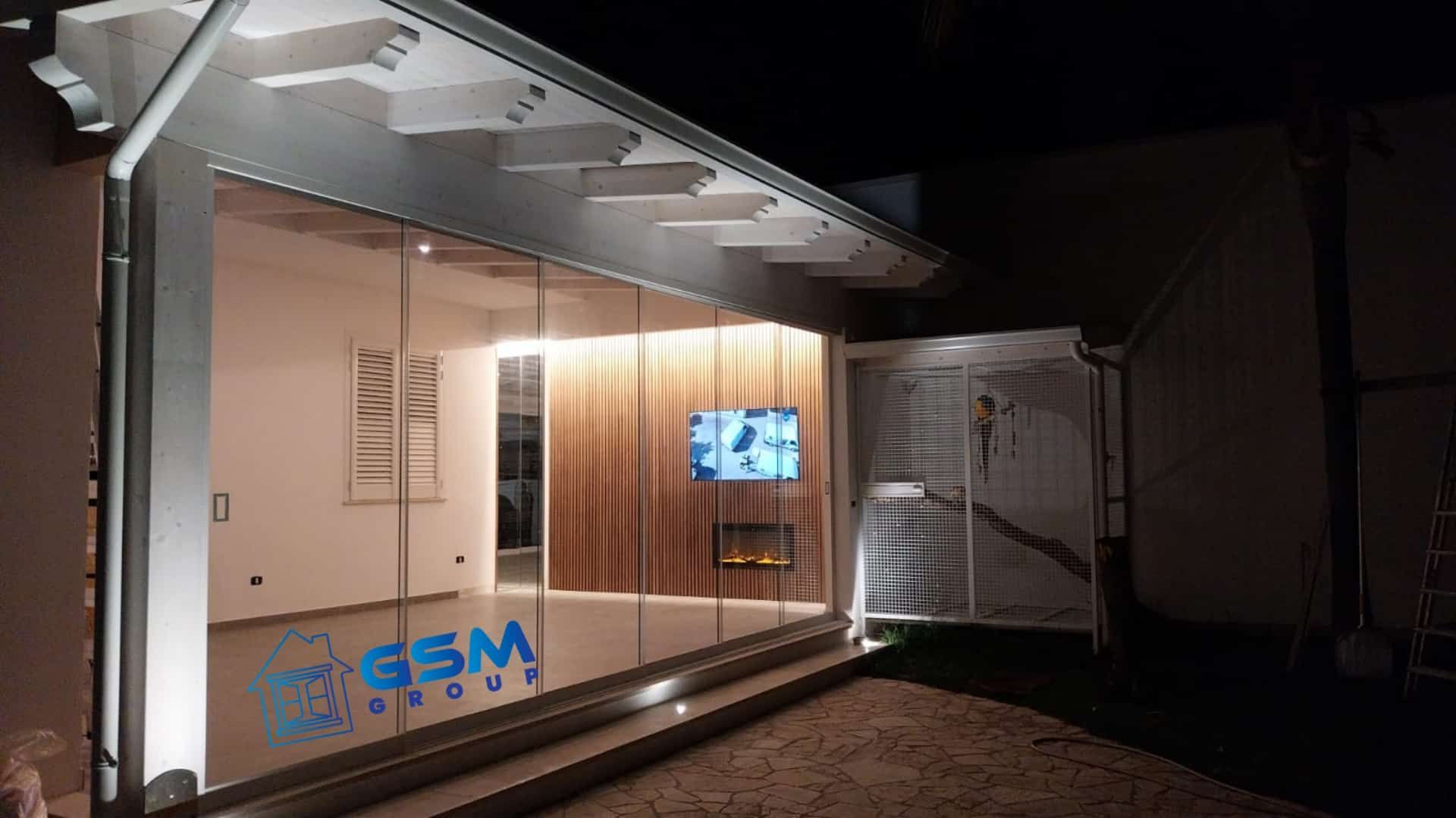 Un moderno patio esterno chiuso da pareti in vetro, caratterizzato da pannelli in legno, una TV a parete e un camino per la sera.