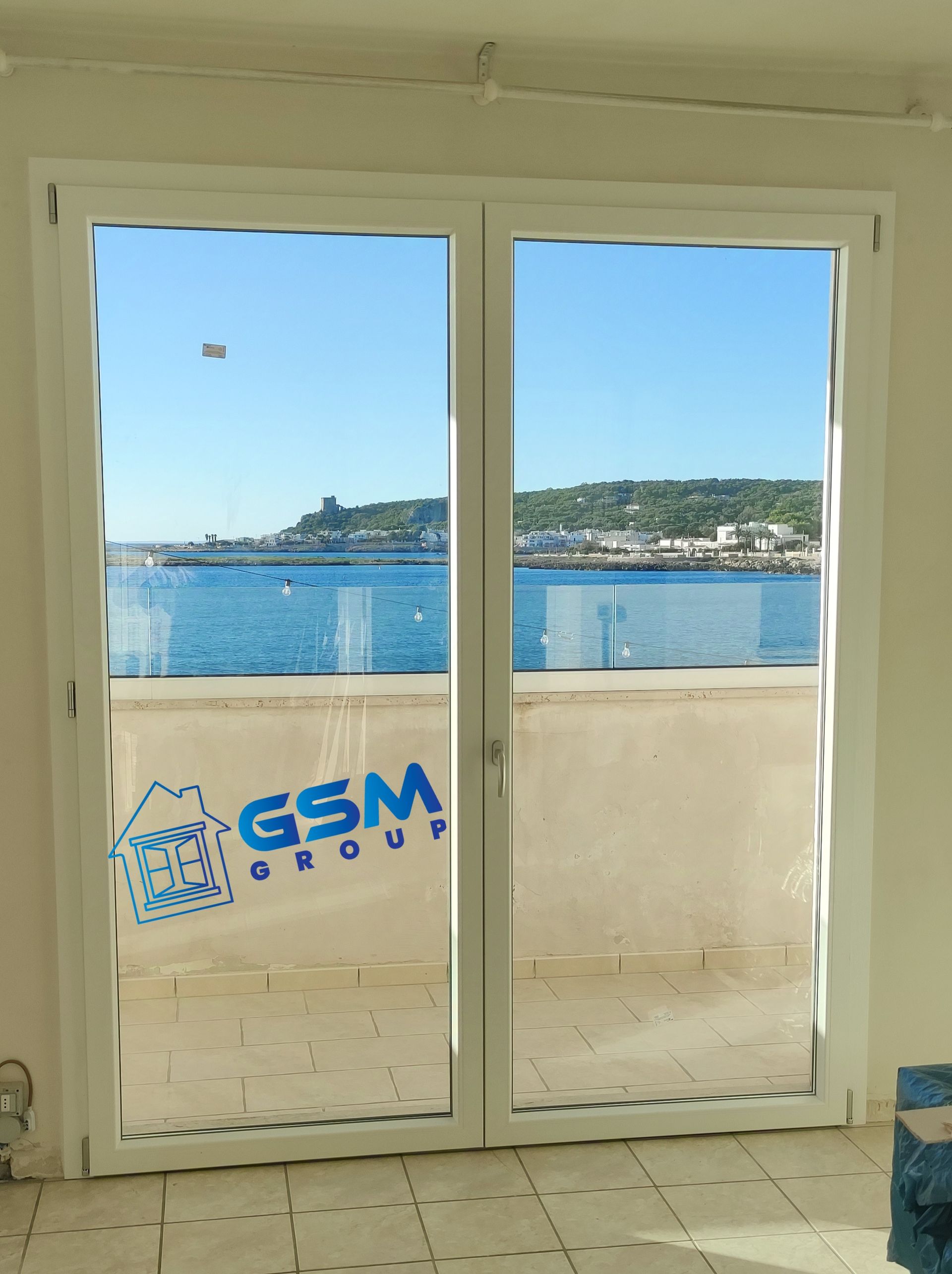 Una porta-finestra in vetro con cornice bianca che si affaccia su un mare azzurro brillante, sulla costa e su un cielo limpido, con il logo del Gruppo GSM.